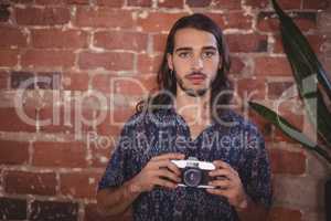 Portrait of confident young photographer holding camera against brick wall