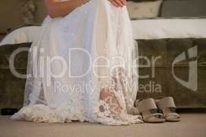 Low section of bride sitting by sandals on bed