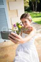 Bride holding bouquet taking selfie with mobile phone while sitting on chair in yard