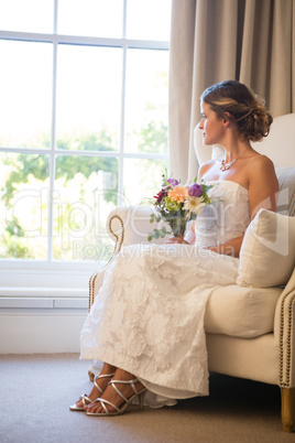 Beautiful bride holding bouquet looking through window while sitting on armchair