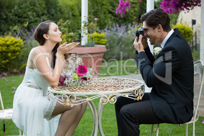 Bridegroom photographing bride blowing kiss while sitting at table in park