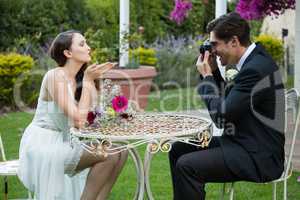 Bridegroom photographing bride blowing kiss while sitting at table in park
