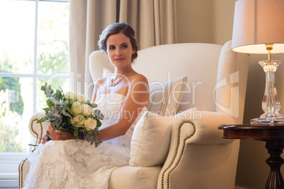 Beautiful bride holding bouquet sitting on armchair at home