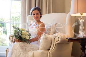 Beautiful bride holding bouquet sitting on armchair at home