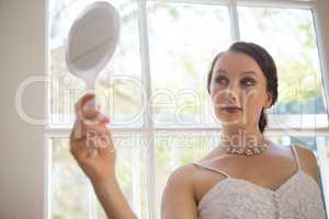 Beautiful bride looking into hand mirror while standing by window at home