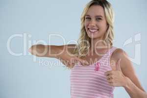 Portrait of smiling woman showing thumbs up while pointing on pink ribbon