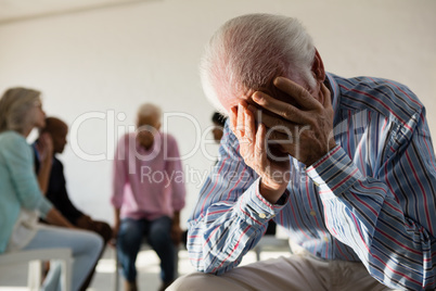 Senior man with head in hand sitting on hair with friends discussing in background