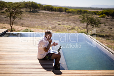 Man using laptop while talking on mobile phone near poolside
