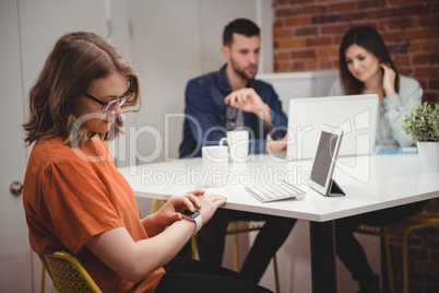 Female executive using smartwatch while colleagues discussing on laptop in background