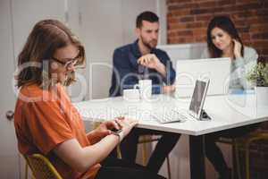 Female executive using smartwatch while colleagues discussing on laptop in background