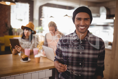 Portrait of smiling young man holding eyeglasses standing against female friends at coffee shop