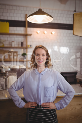 Portrait of smiling young waitress standing with hands on hip against counter