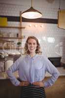 Portrait of smiling young waitress standing with hands on hip against counter
