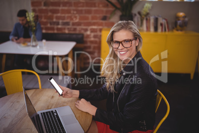 Smiling young blond woman holding smartphone while sitting with laptop at coffee shop