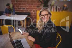 Smiling young blond woman holding smartphone while sitting with laptop at coffee shop