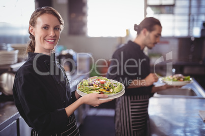 Portrait of smiling young waitress holding fresh salad against waiter at commercial kitchen