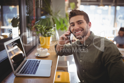 Portrait of smiling young man talking smartphone while sitting by laptop