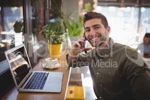 Portrait of smiling young man talking smartphone while sitting by laptop