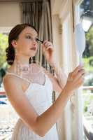 Beautiful bride applying mascara while looking into hand mirror by window