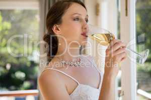 Bride looking through window while drinking champagne at home