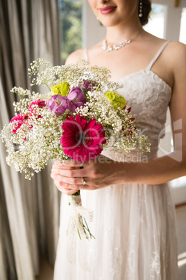 Midsection of smiling bride in wedding dress holding bouquet at home