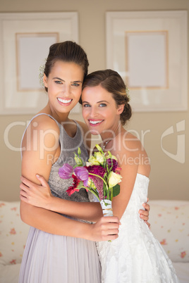 Portrait of happy bride and bridesmaid with bouquet standing in bedroom