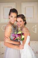Portrait of happy bride and bridesmaid with bouquet standing in bedroom