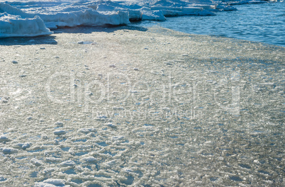 Melting ice chunks by water and frozen shore.