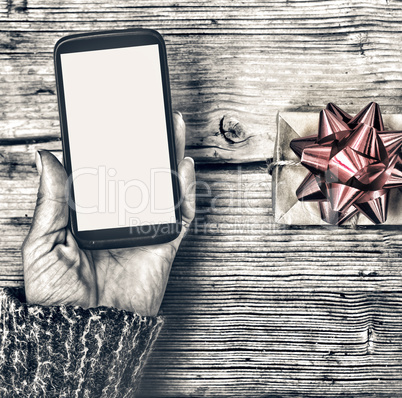 Closeup of a smartphone in the hands of a woman and a gift box with a red bow on a wooden table.