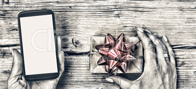 Closeup of a smartphone in the hands of a woman and a gift box with a red bow on a wooden table.