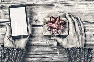 Closeup of a smartphone in the hands of a woman and a gift box with a red bow on a wooden table.