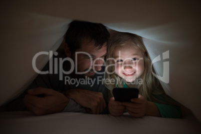 Father and daughter lying under blanket and using mobilephone