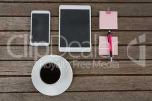 Black coffee, mobile phone, digital tablet, pen and sticky note on wooden plank