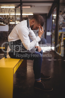 Side view of upset young waiter sitting on yellow bench with headache