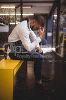 Side view of upset young waiter sitting on yellow bench with headache