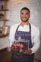 Portrait of smiling young waiter holding cake on glass cakestand