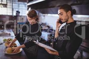 Young waiter talking on smartphone while waitress preparing food in commercial kitchen