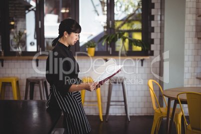 Side view of young waitress reading menu while leaning on table