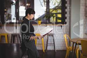 Side view of young waitress reading menu while leaning on table