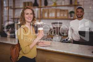 Portrait of smiling young female customer holding disposable coffee cup at counter against waiter