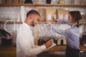 Young male owner writing on clipboard while waitress arranging products