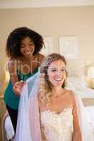 Beautician adjusting veil of bride hair while sitting on bed at home