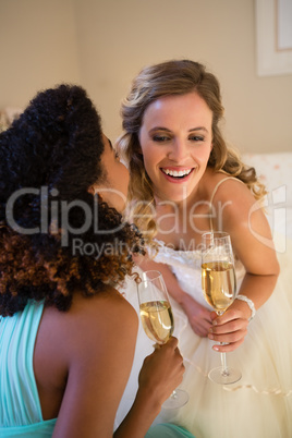 Bridesmaid whispering into bride ear while having champagne in bedroom