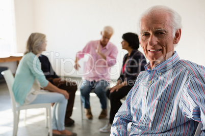 Portrait of senior man sitting on chair with friends discussing in background