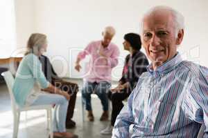 Portrait of senior man sitting on chair with friends discussing in background