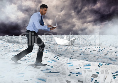 Businessman on laptop in sea of documents under sky clouds with paper boats
