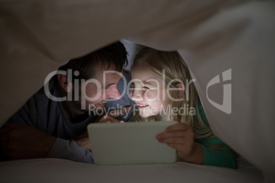 Father and daughter lying under blanket and using digital tablet