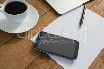 Laptop, mobile phone, black coffee, pen and paper on wooden plank