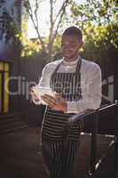 Smiling young waiter using digital tablet while leaning on railing