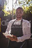 Portrait of smiling young waiter using digital tablet while leaning on railing